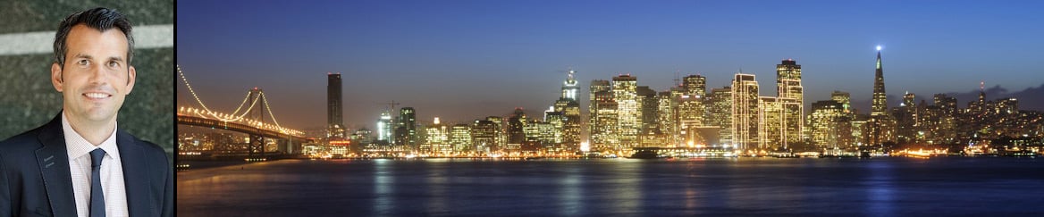 Smiling man in a suit beside a lit-up San Francisco skyline at dusk with the Bay Bridge.